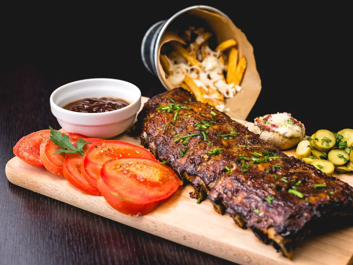A wooden board holding barbecue pork ribs, sliced tomato, pickles and a paper cone of fries.